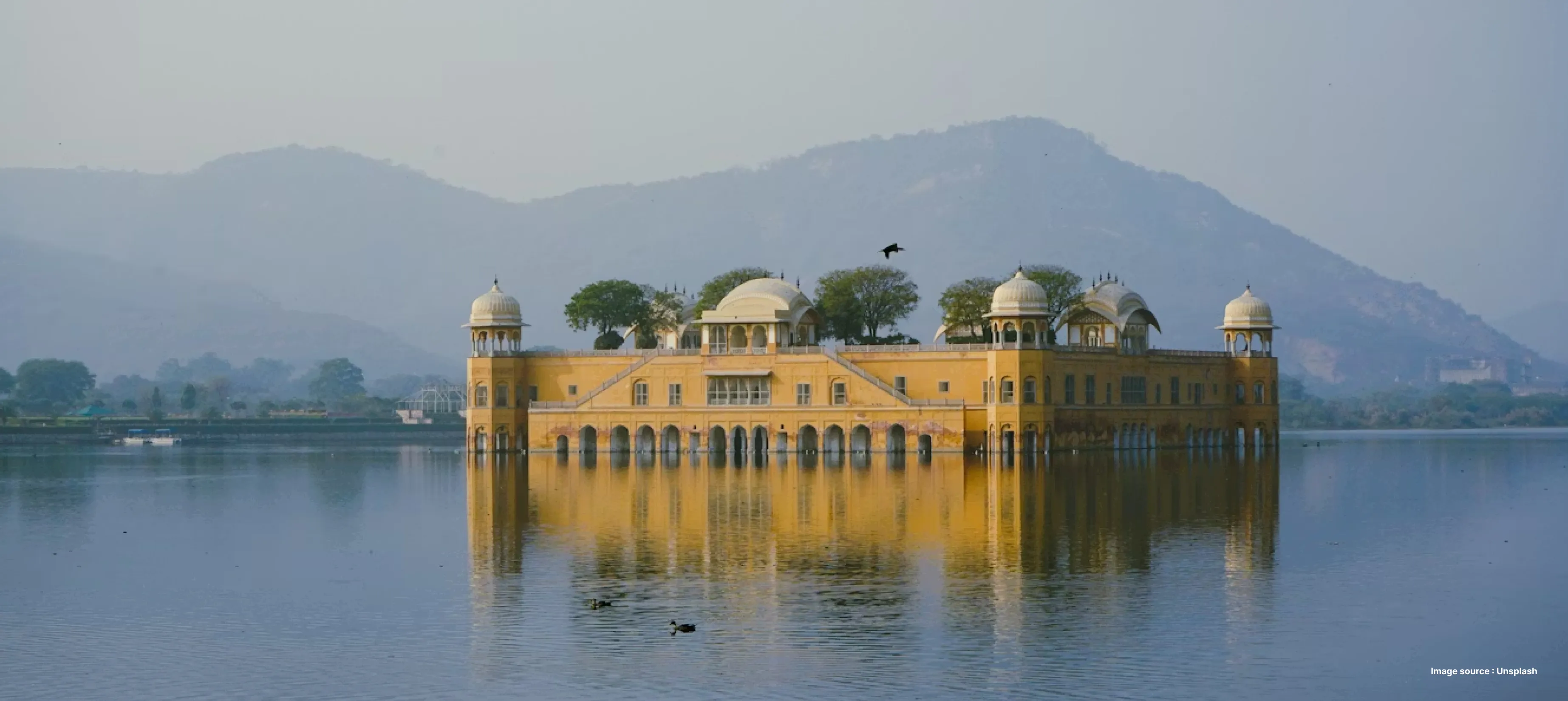 a closer view of the jal mahal in jaipur - its yellow palace building seems to float in the middle of the lake with green hills in the background