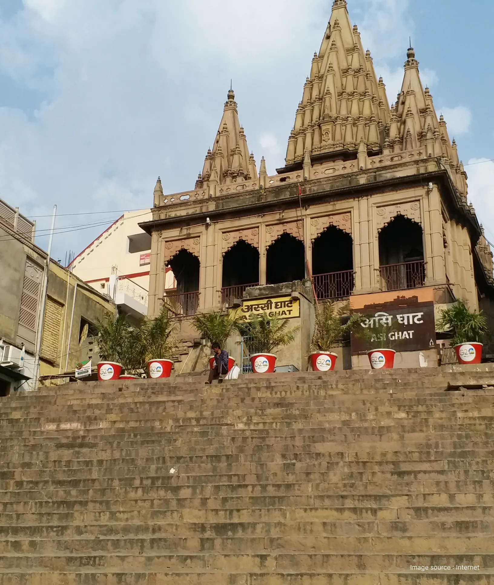 A view of the Assi Ghat entrance in Varanasi featuring traditional Hindu temple spires, stone stairs, and decorative potted plants with G20 branding.