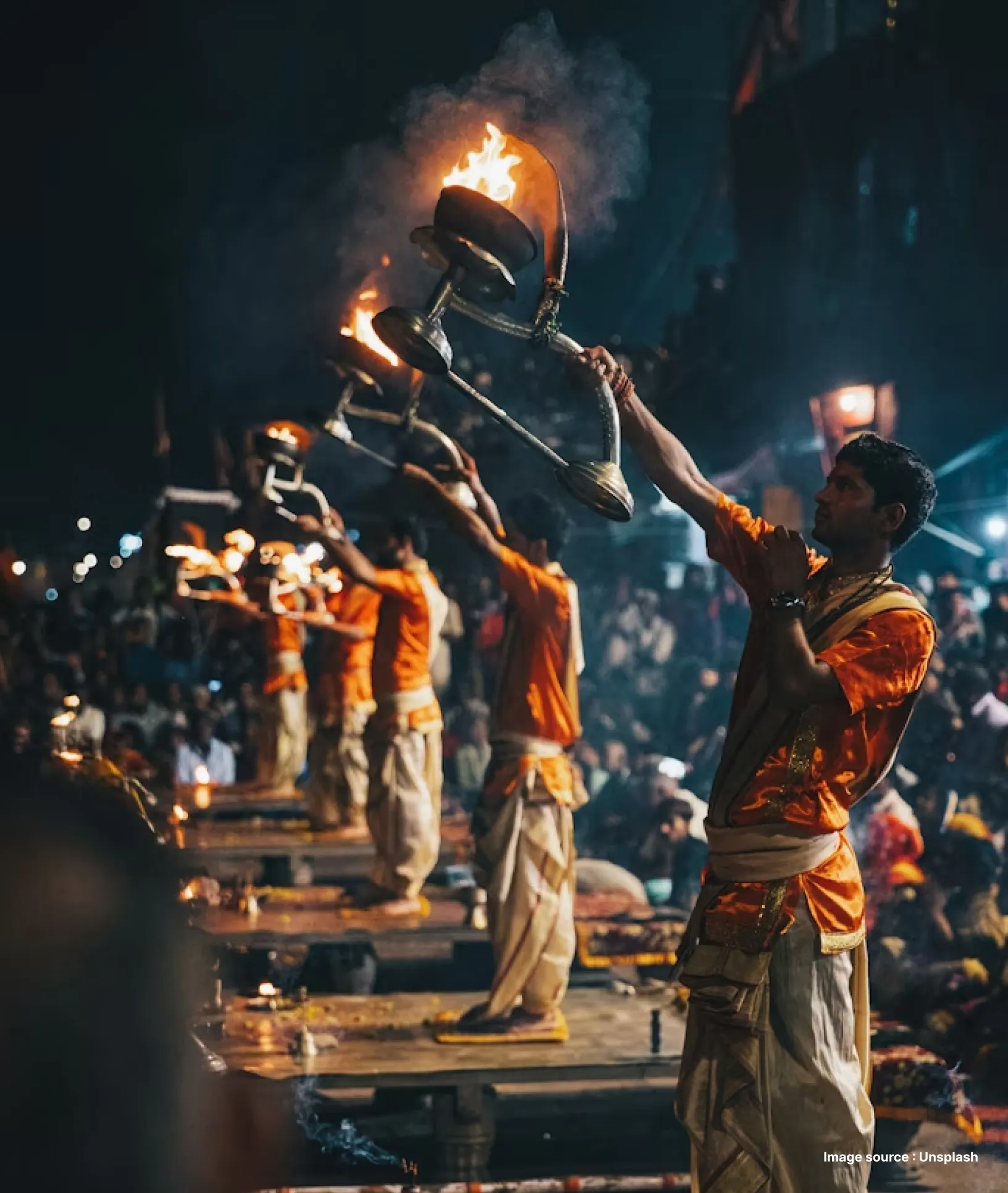 a group of priests dressed in orange perform worship with large lamps in synchronisation at night