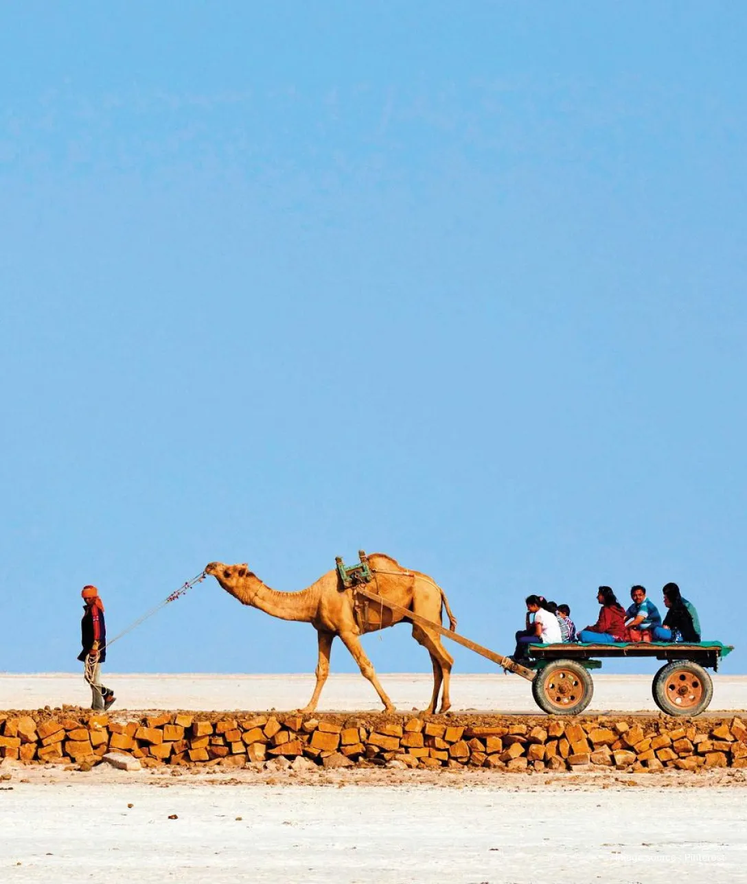 side view of a man guiding a camel cart with tourists ahead in the white desert