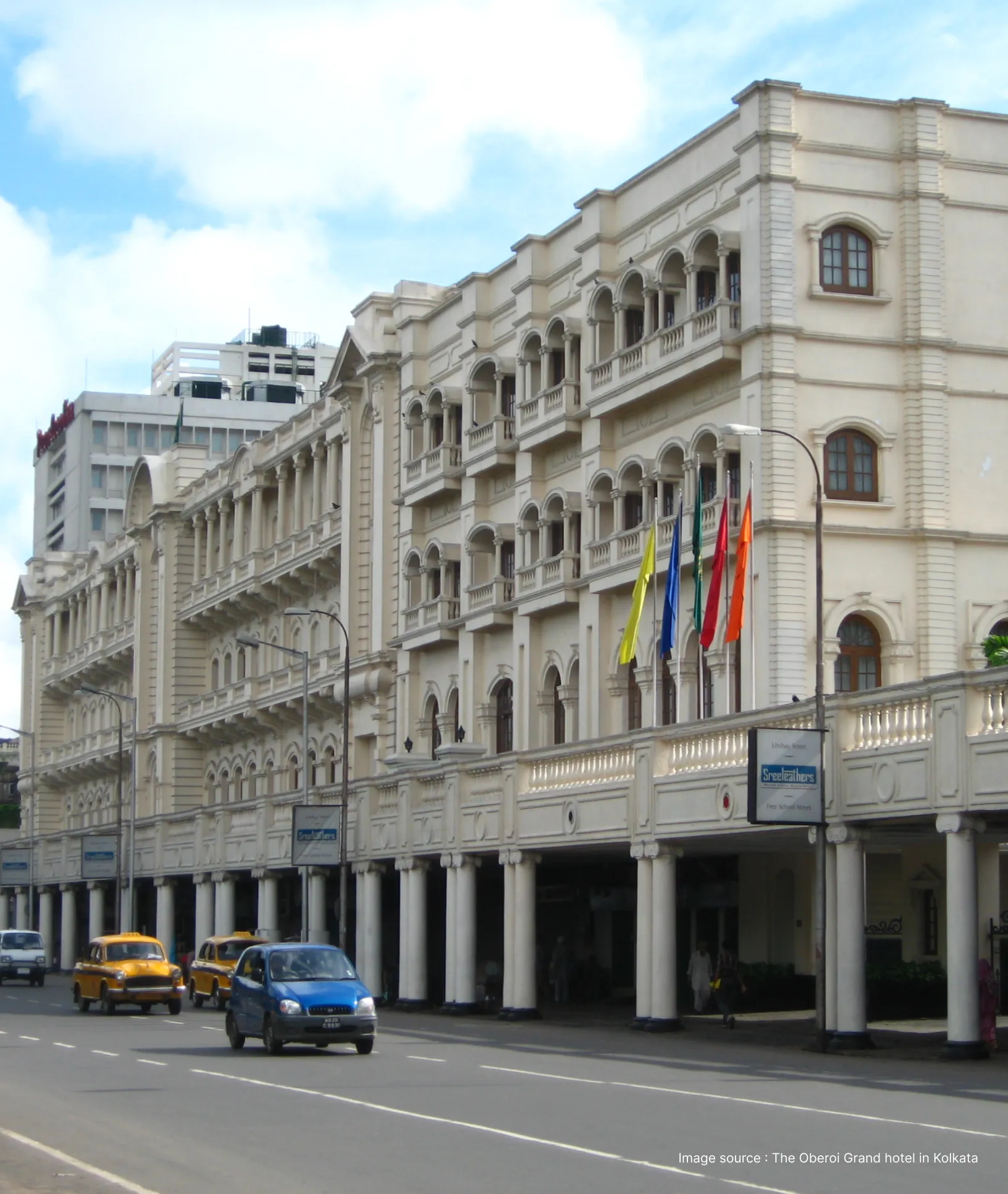 outside view of the all-white oberoi grand building with a blue and yellow taxis passing in front of it