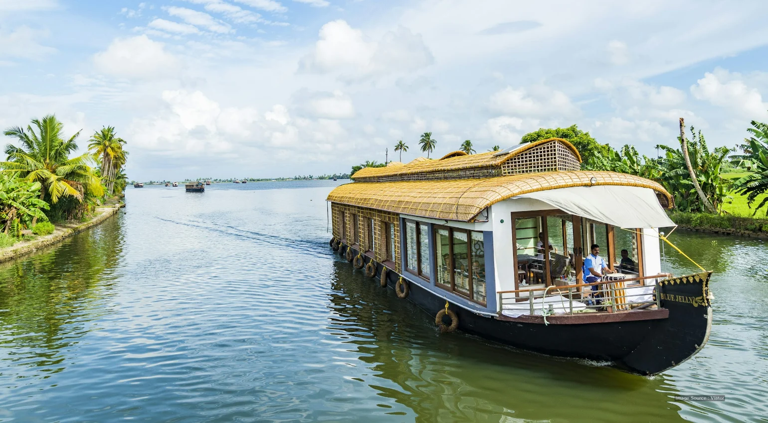 A traditional houseboat cruising through the lush green backwaters, lined with palm trees, showcasing one of the scenic travel destinations in Kerala.