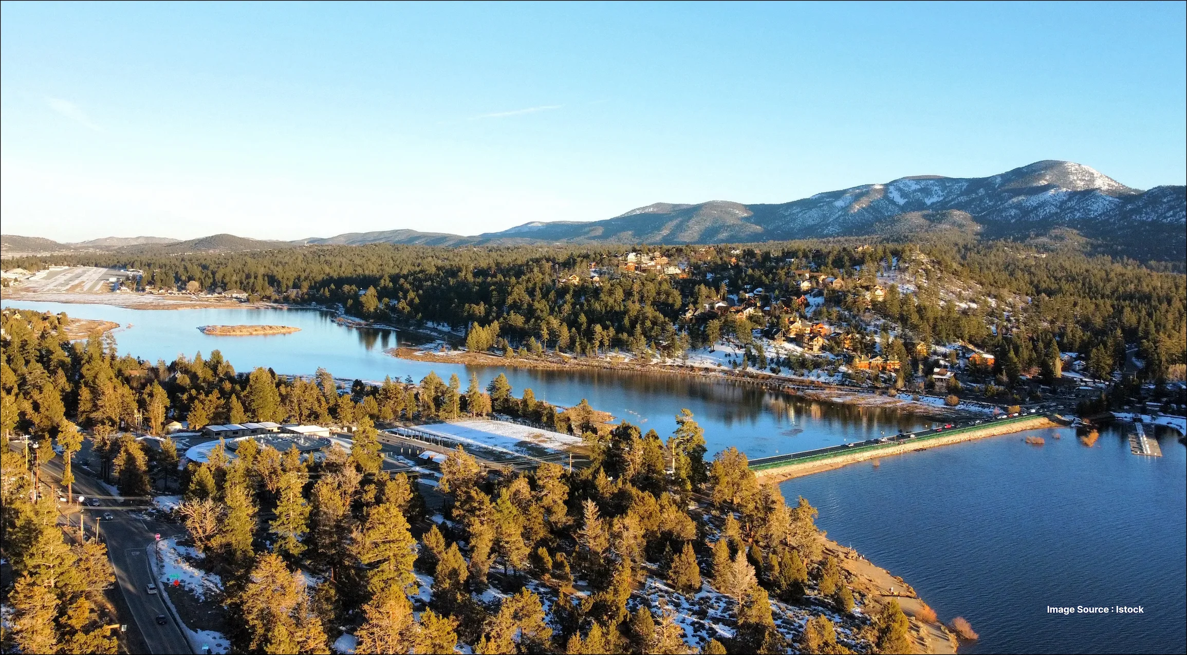 Aerial view of a mountain lake surrounded by pine forest, with snow-dusted hills, lakeside cabins, and distant mountains under a clear blue sky.