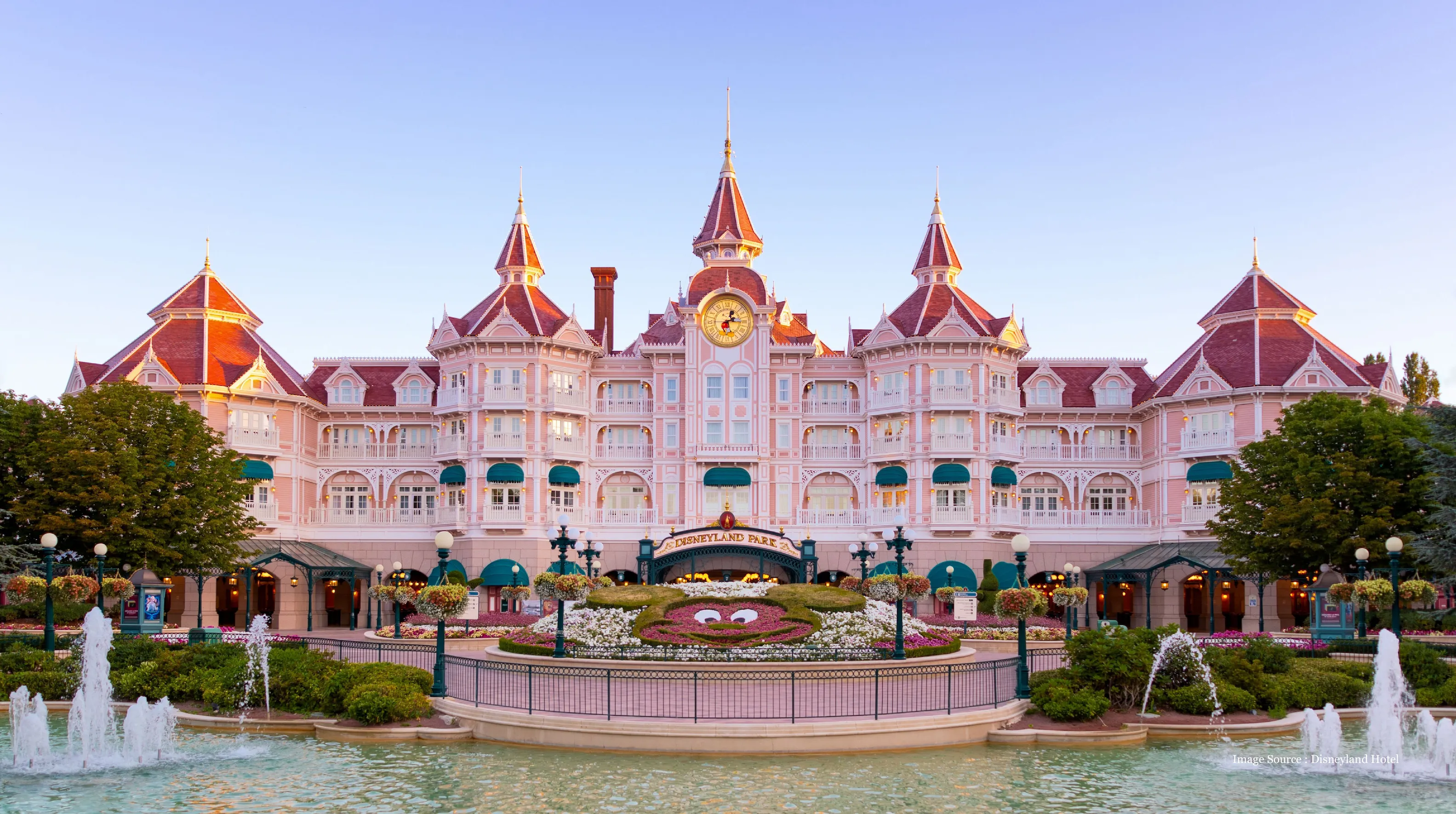 Front view of the pink Victorian-style Disneyland Hotel with a central clock tower, lush gardens, fountains, and the entrance to Disneyland Park.