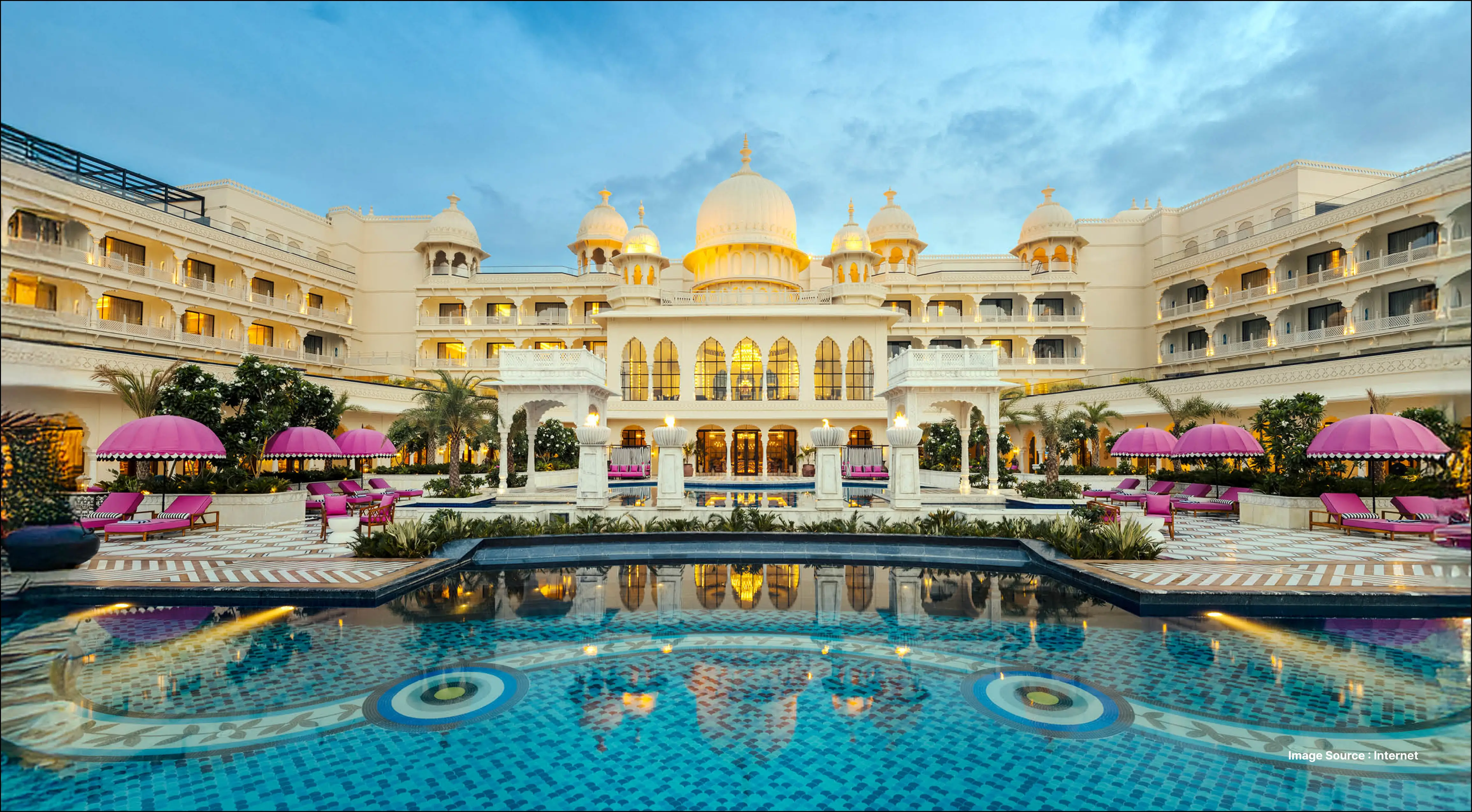 Luxurious palace-style hotel in Udaipur with grand domes, illuminated arches, and a reflecting pool surrounded by pink lounge umbrellas at dusk