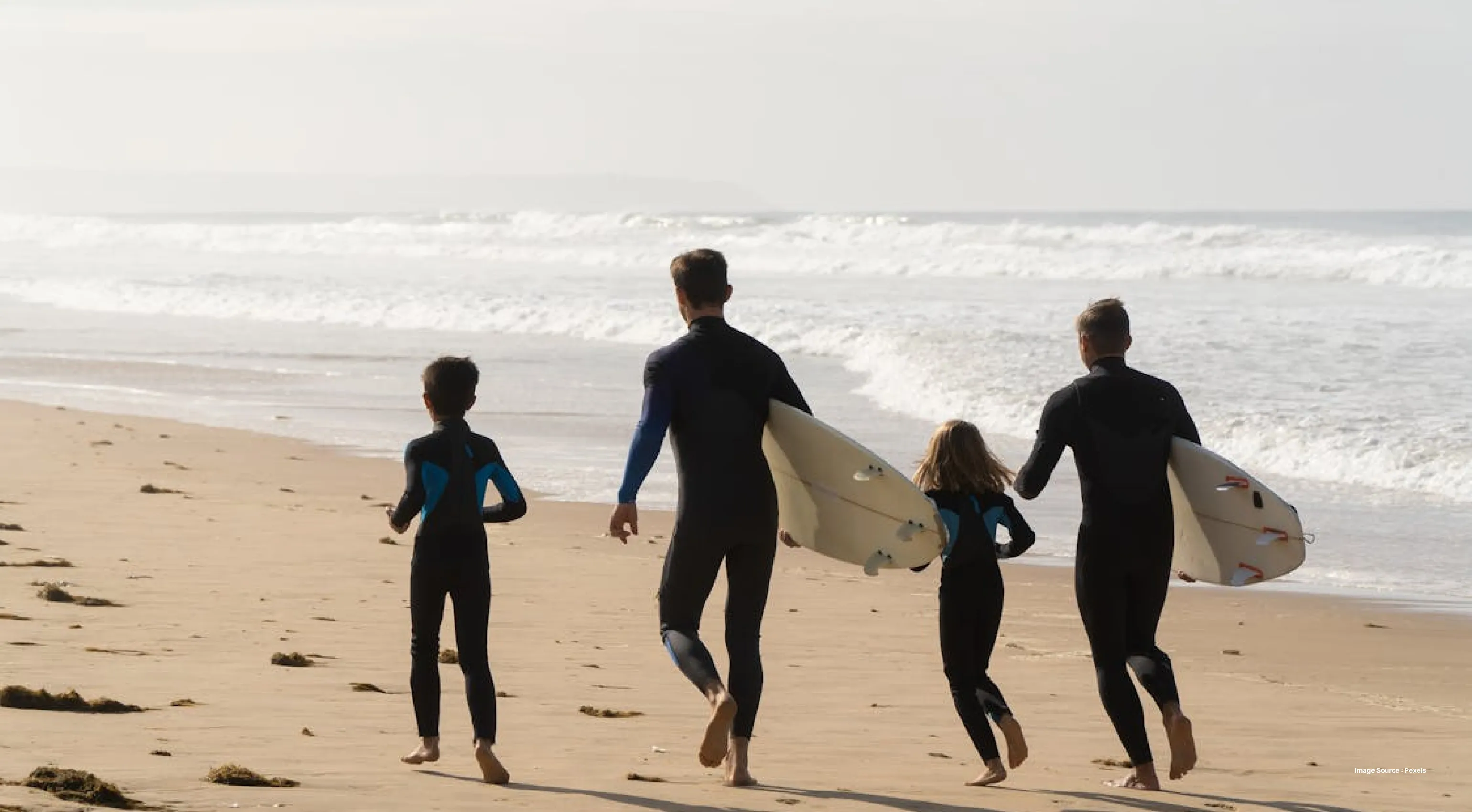a familywith two kids dressed in surfing suits with surfboards, walking on the beach