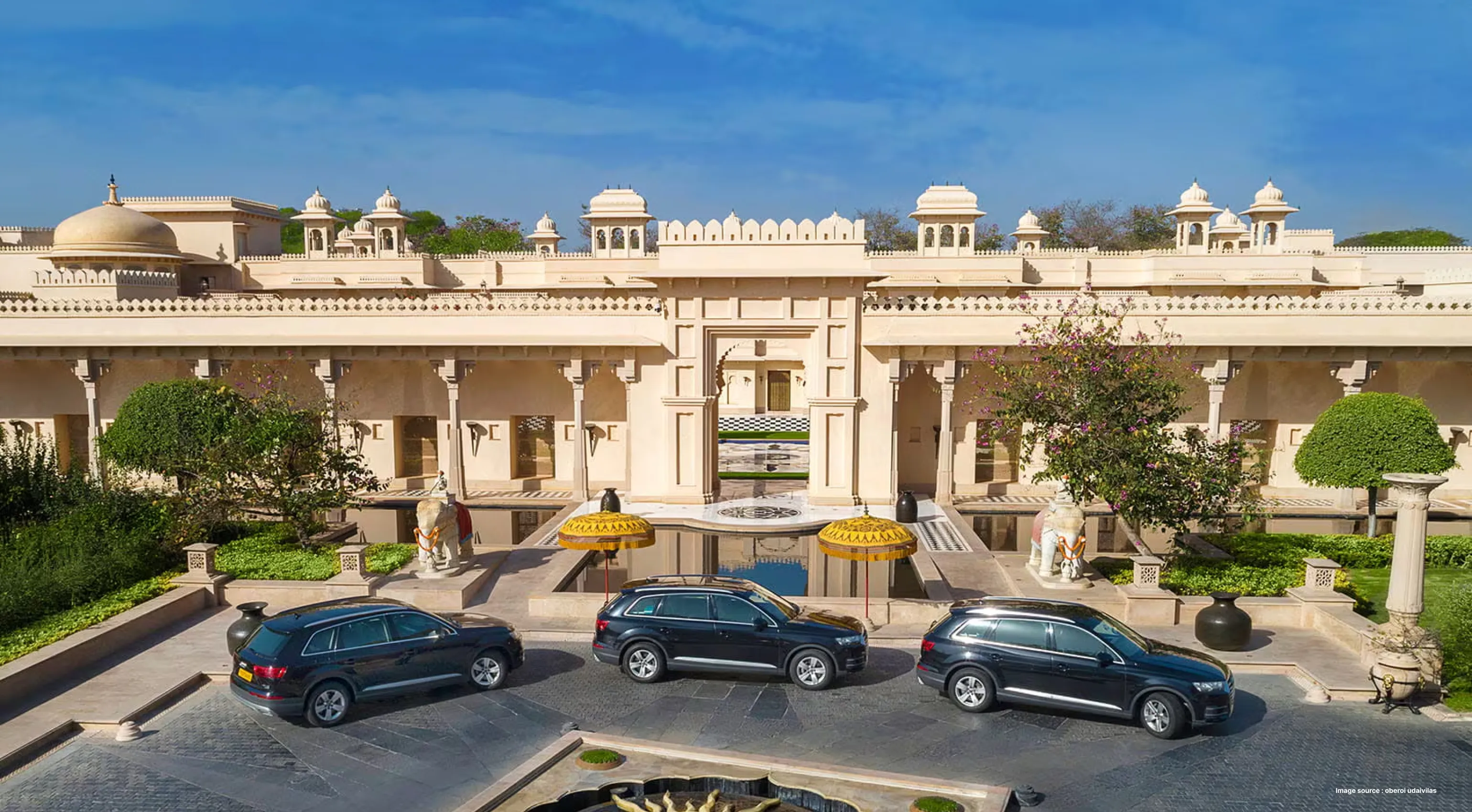 three luxury vehicles parked in front of a luxury hotel with indian architecture and decorative trees