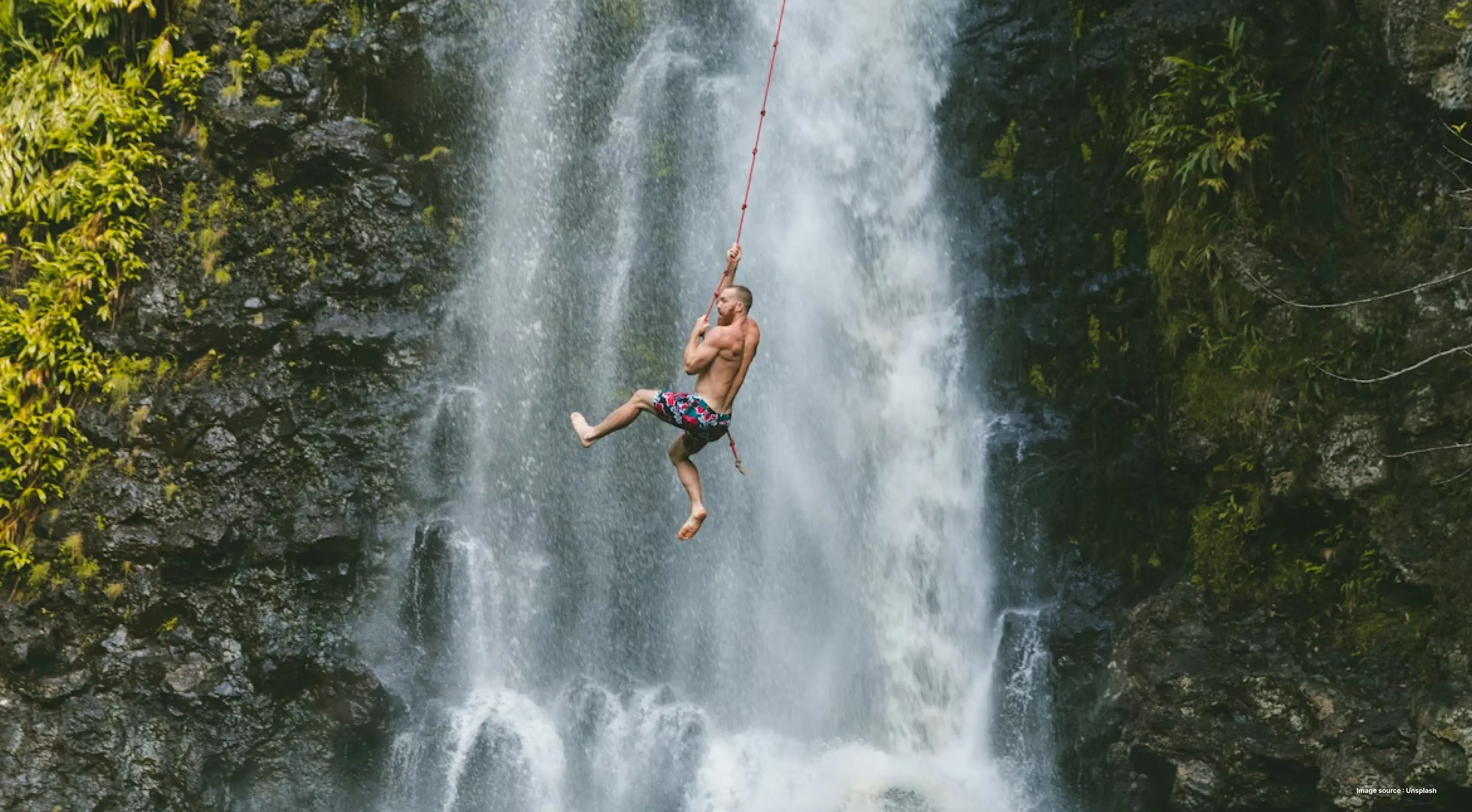 a white man in shorts swinging on a rope across a waterfall