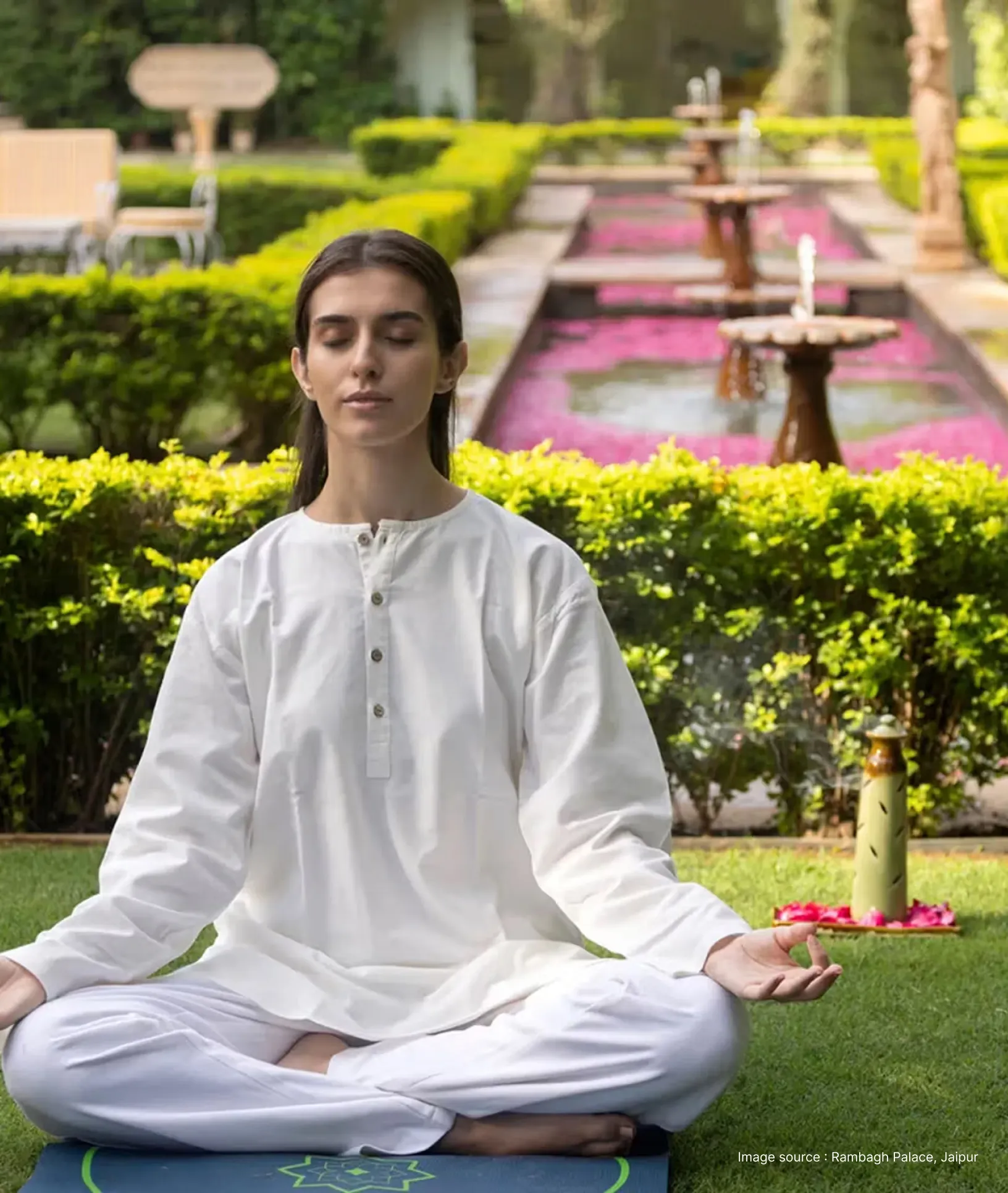 a white woman dressed in white kurta seated in a serene garden with fountains practicing yoga