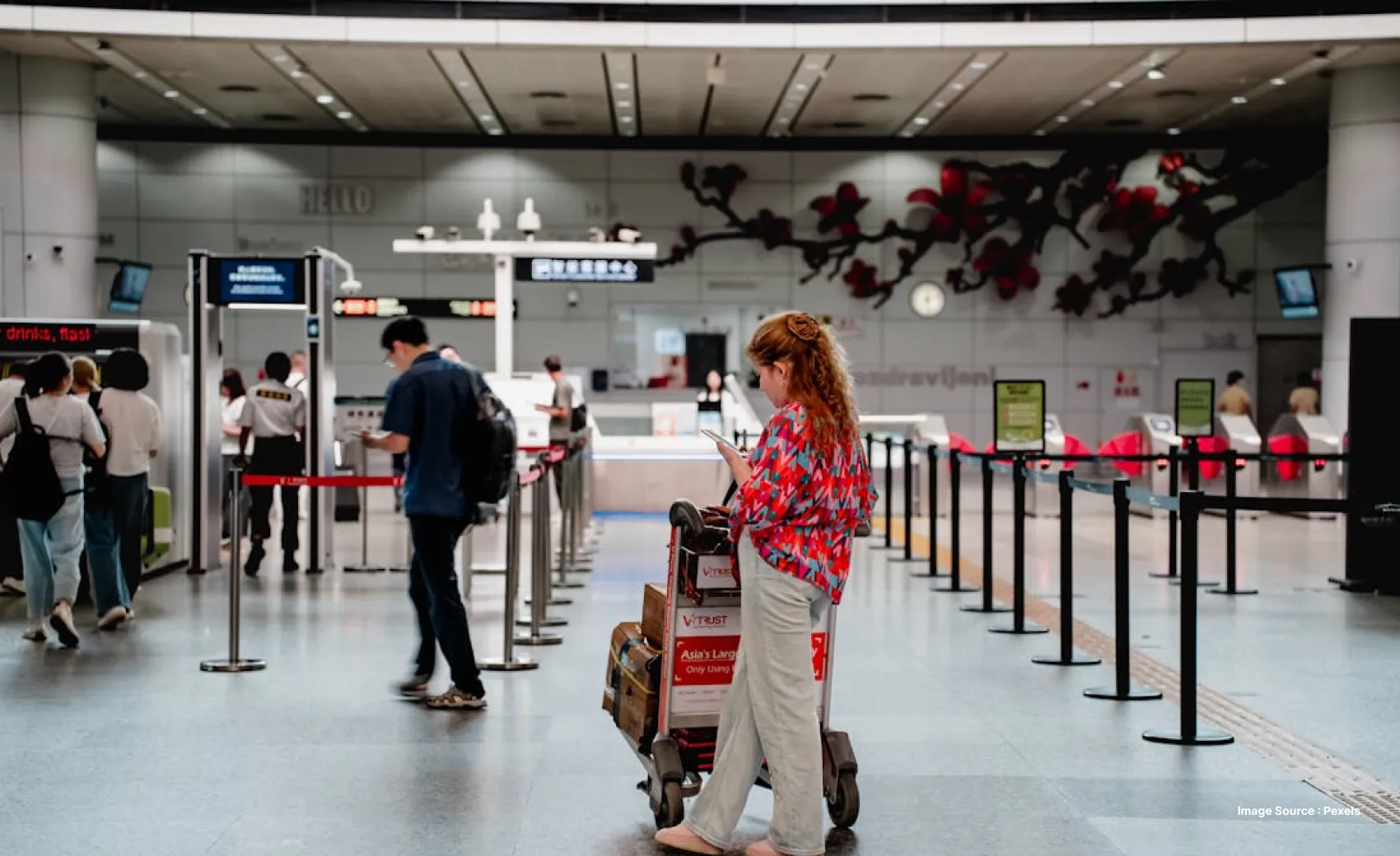 a lady in a colorful top looks at her phone while waiting with a luggage trolley at airport security