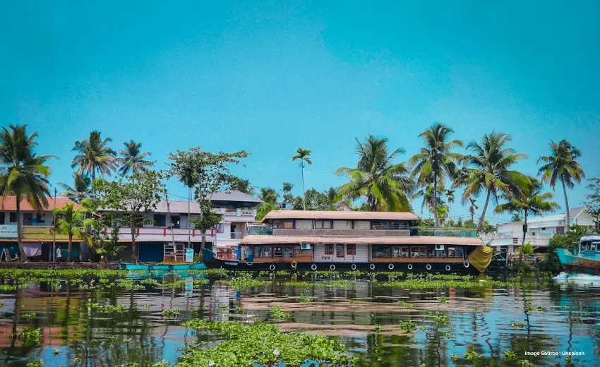 a kerala houseboat parked on the shore of a backwater with palm trees and building in the background