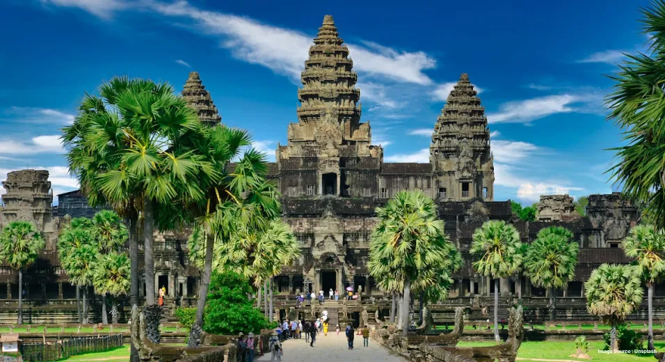 the ancient angkor wat temple with tourists walking up to its entrance and the path flanked by palm trees