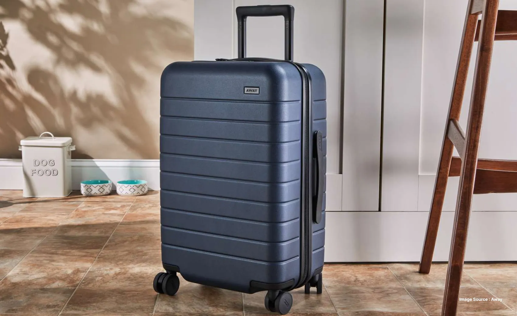A dark blue suitcase at a home next to a white cupboard and a wooden stool