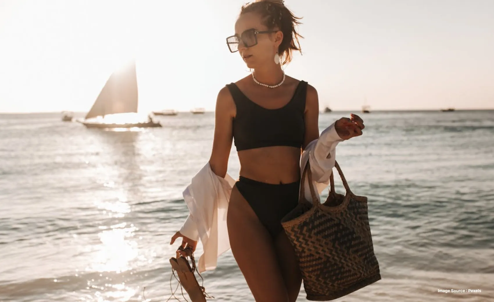 a woman in a black bikini set with a beach bag walking on a beach