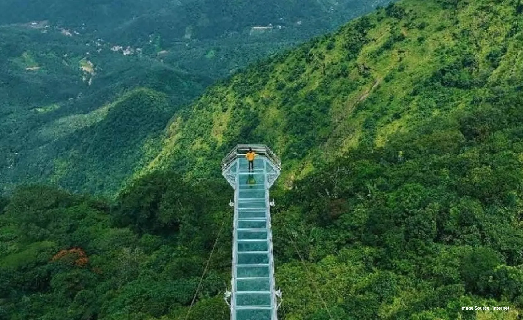 Aerial view of lush green mountains and a glass bridge with a person standing at the edge of it