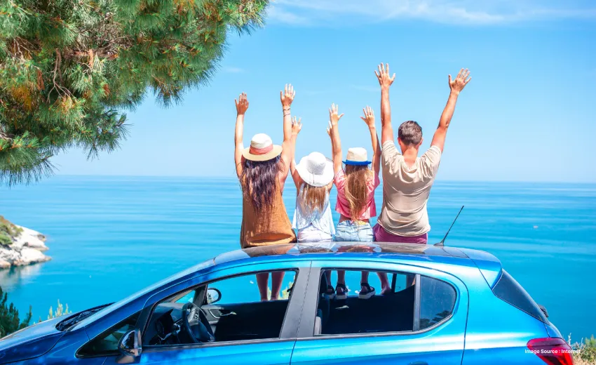 a family of four sitting on top of a blue car with hands up in joy in front of a beach with hats on