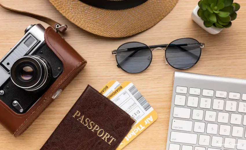 Travel essentials laid out on a wooden desk including a vintage camera, sunglasses, passport with boarding passes