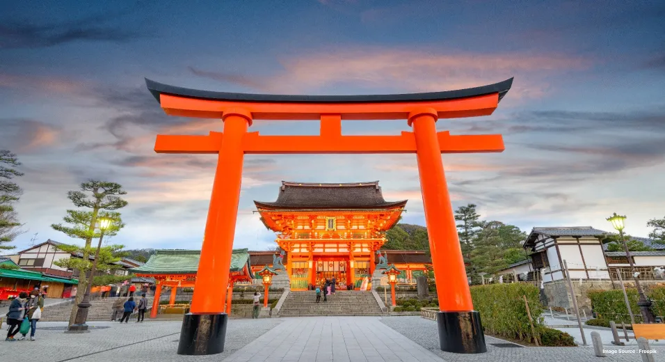 an orange japanese gate leading up to a japanese temple where devotees are walking in at sunset