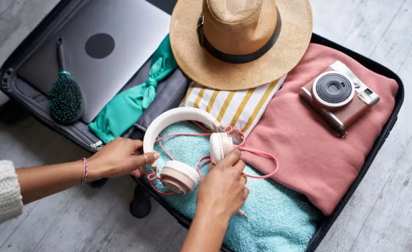 An open suitcase, showing items like clothes, hairbrush, camera, laptop, and hat being packed for a vacation