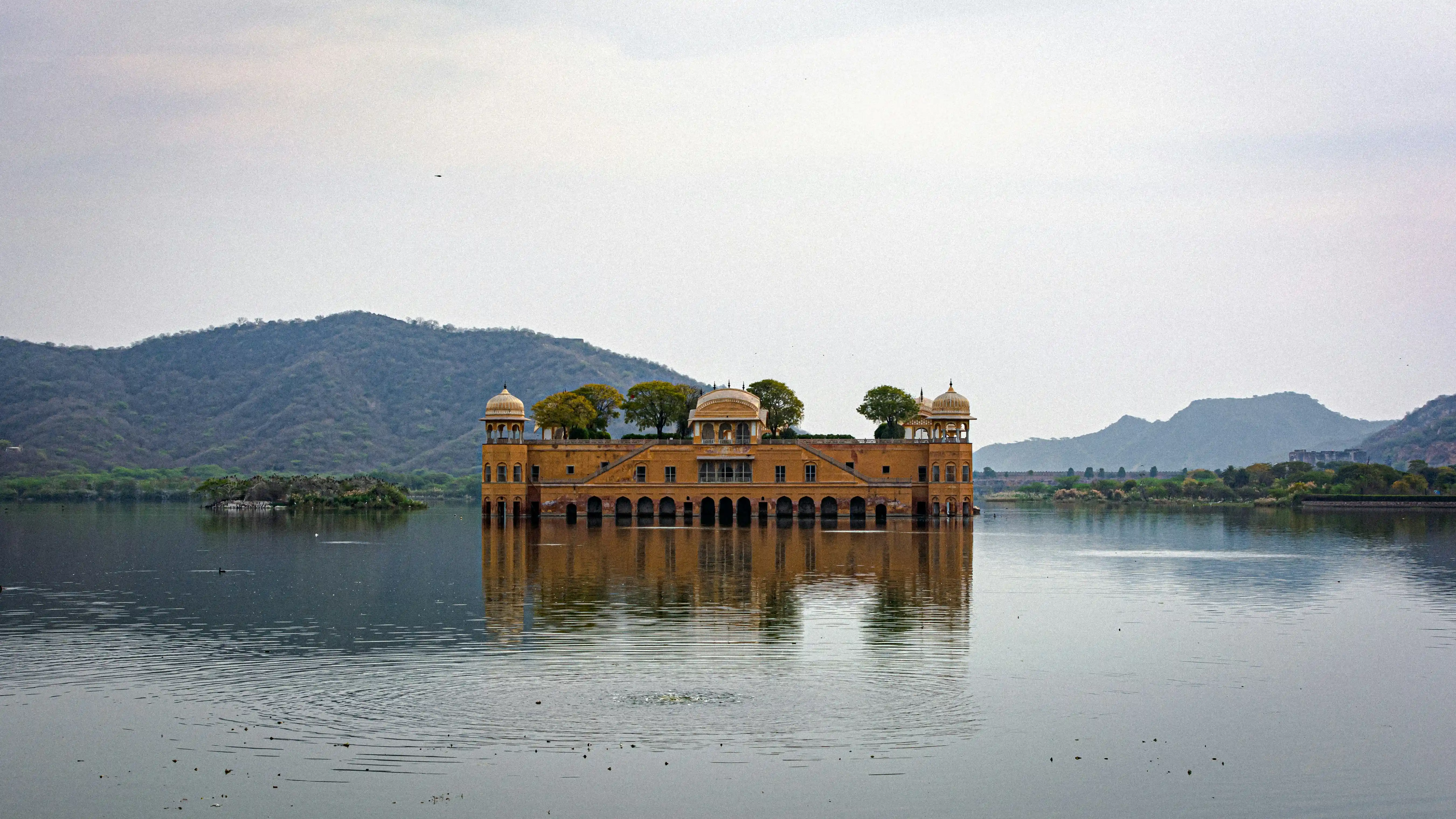 a yellow palace in the middle of the mansagar lake with small green hills in the background