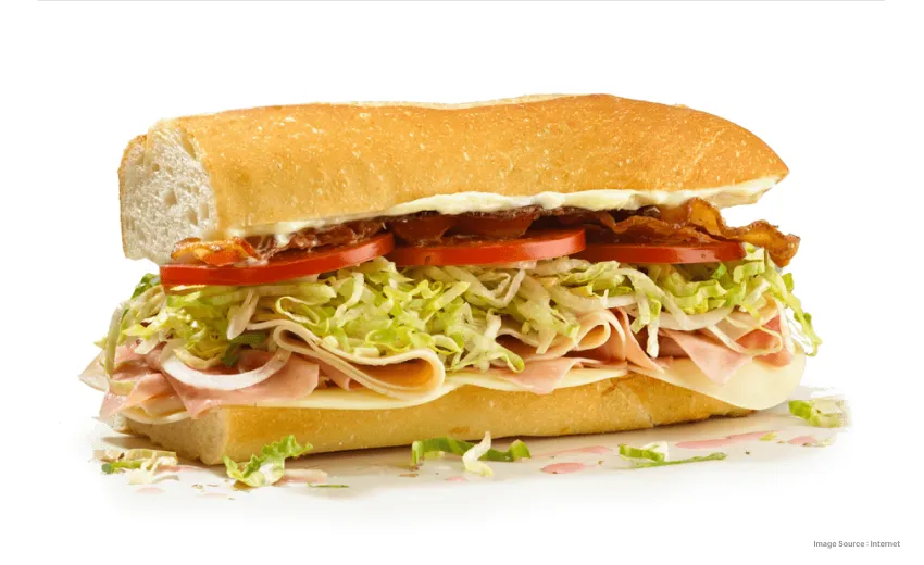 Close-up of a deli-style sub sandwich filled with sliced meat, shredded lettuce, tomatoes, onions, and bacon on a long bread roll.