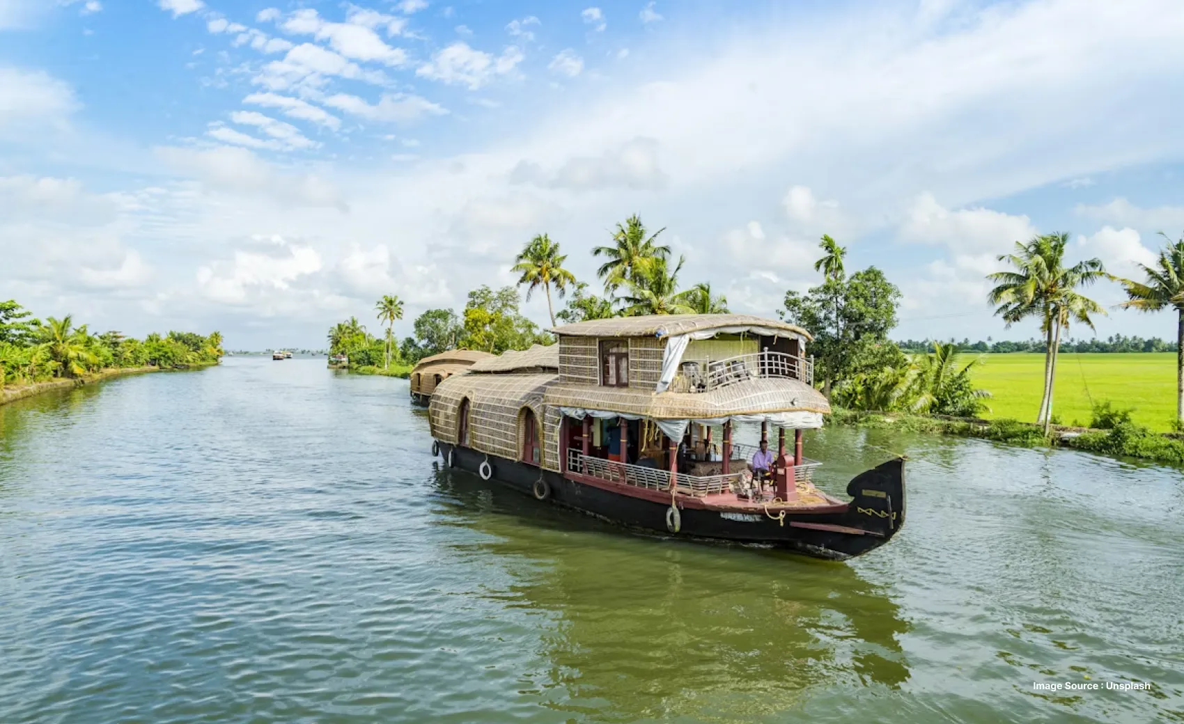 a traditional wooden houseboat cruises ahead on backwaters in kerala with a man guiding it