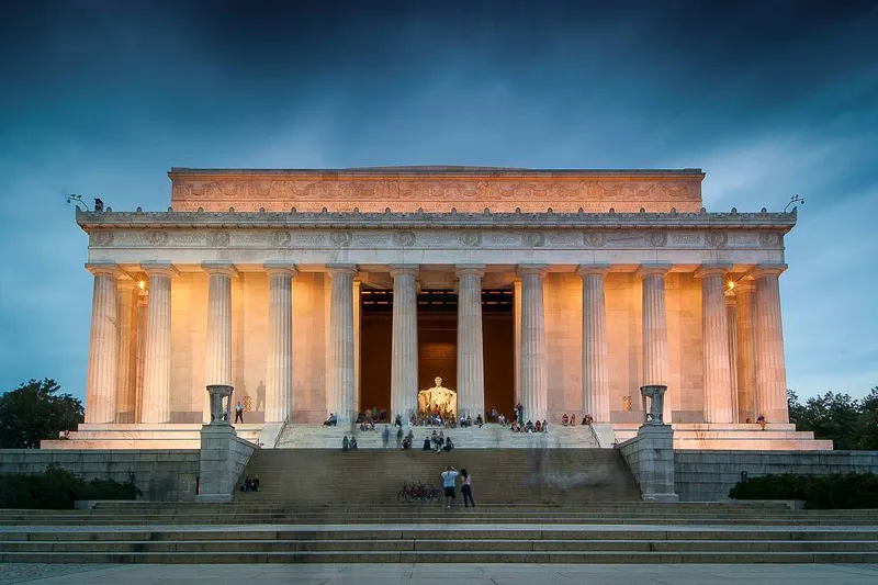 the white building of lincoln memorial lit up at dusk with tourists entering 