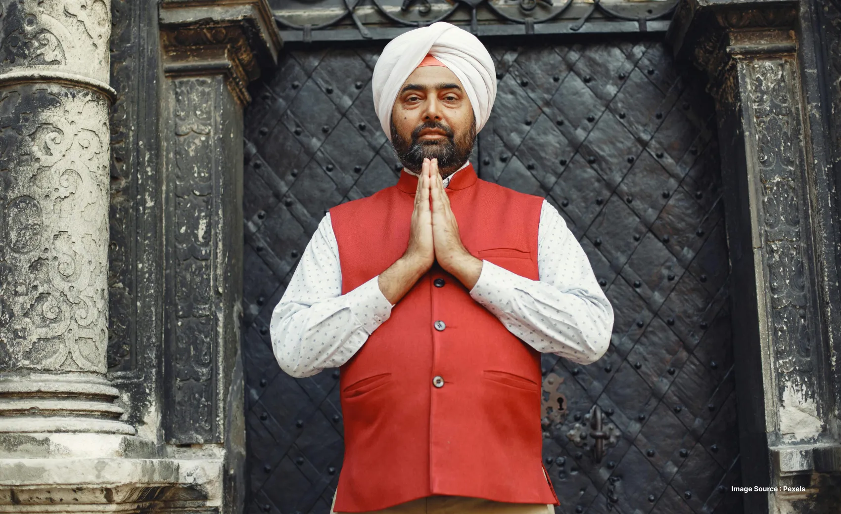 an indian man in red turban putting palms together to say sat sri akaal or namaste with a rustic background