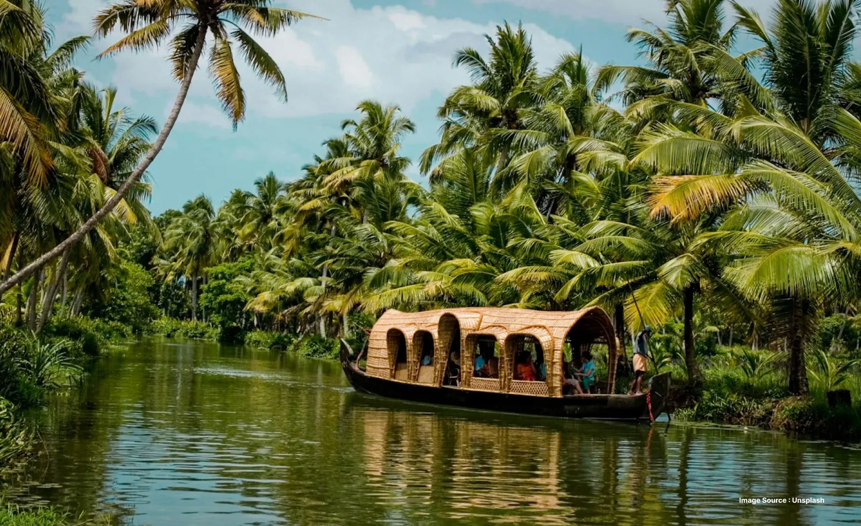 a traditional wooden houseboat with tourists in them by shore flanked by palm trees floating down the backwaters