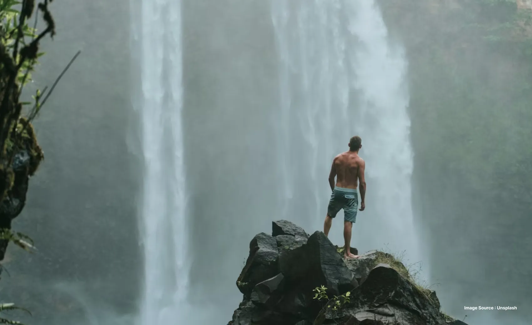 a white man in shorts stands on a huge rock looking ahead at a waterfall