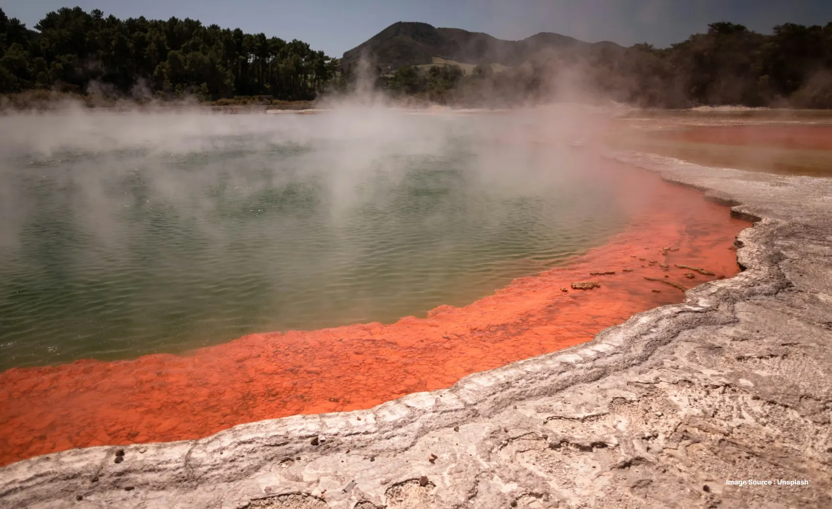 a natural spring with red deposits by the shore as steam rises from the water body, surrounded by green hills