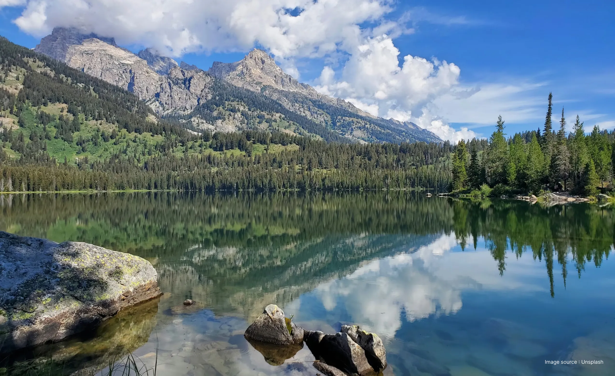 a view of the taggart lake during daytime, surrounded with grey cliffs of the tetons and green trees