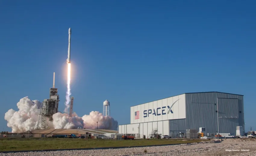 a rocket blasts off leaving behind fire and smoke next to the spacex building