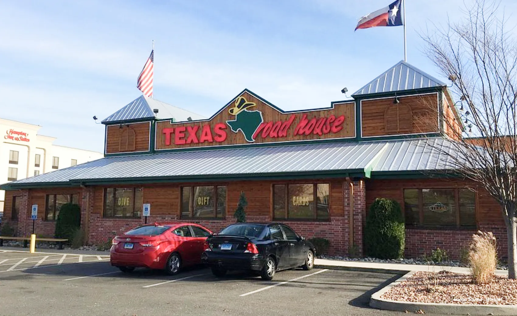 a branch of texas roadhouse restaurant with two cars parked in the front and flags hoisted on top