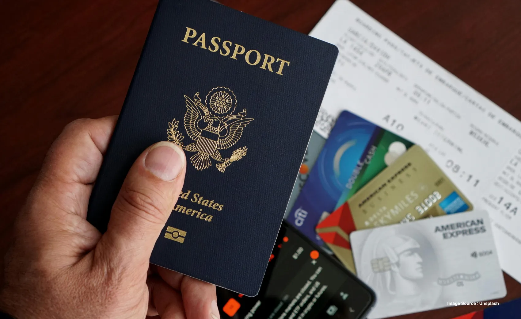 a person's hand holding a usa passport with some credit cards and a boarding pass lying on the table below