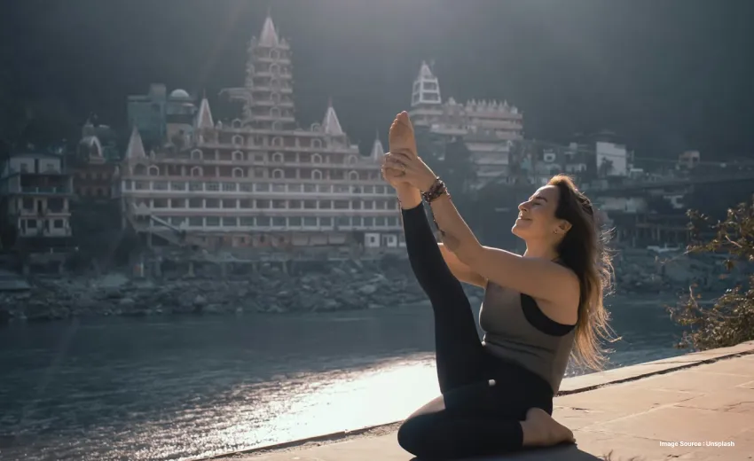 a white woman practicing a yoga pose in front of the ganga river in rishikesh in the morning