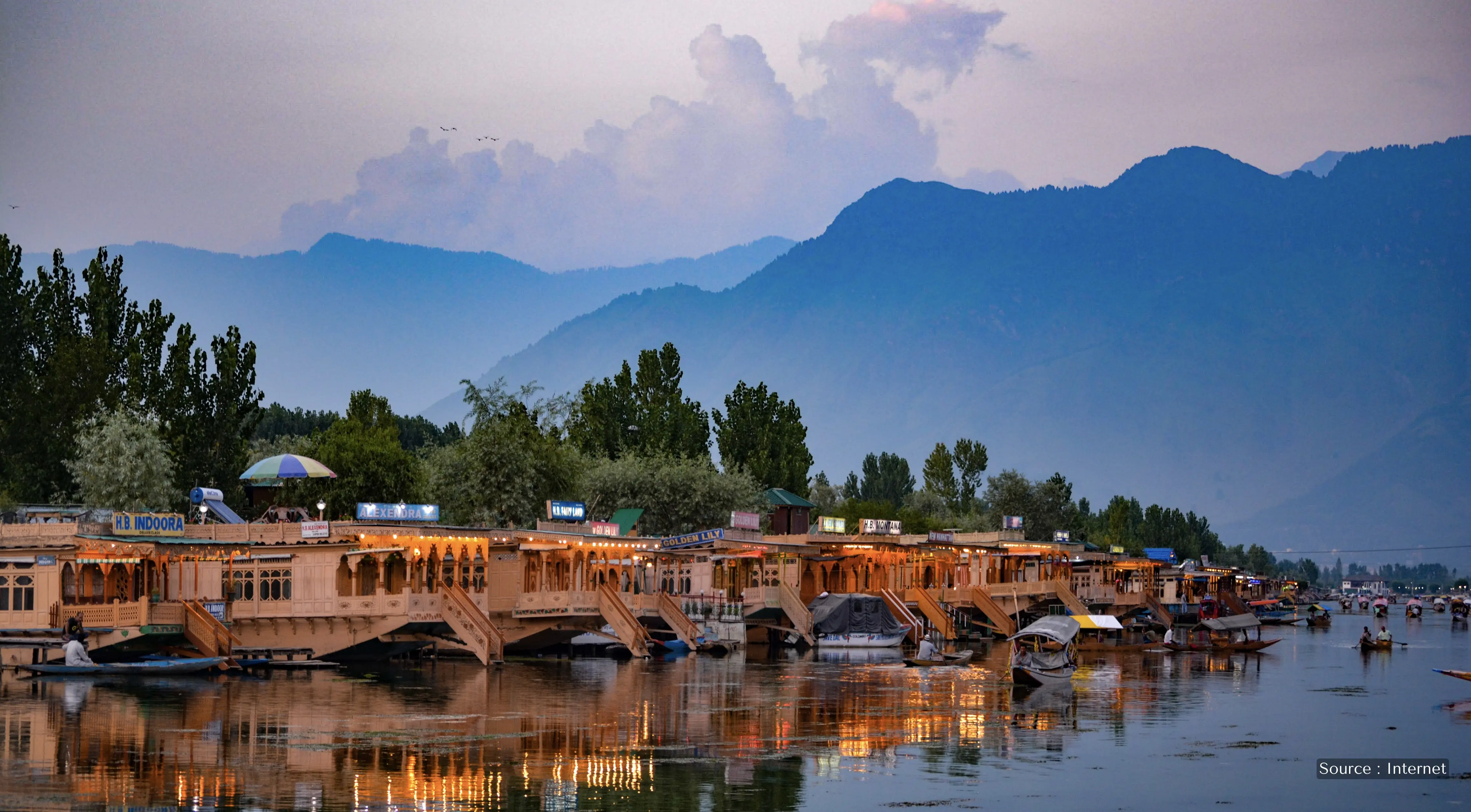 Traditional wooden houseboats illuminated at dusk on Dal Lake, Srinagar, Kashmir, with Himalayan mountains in the background.