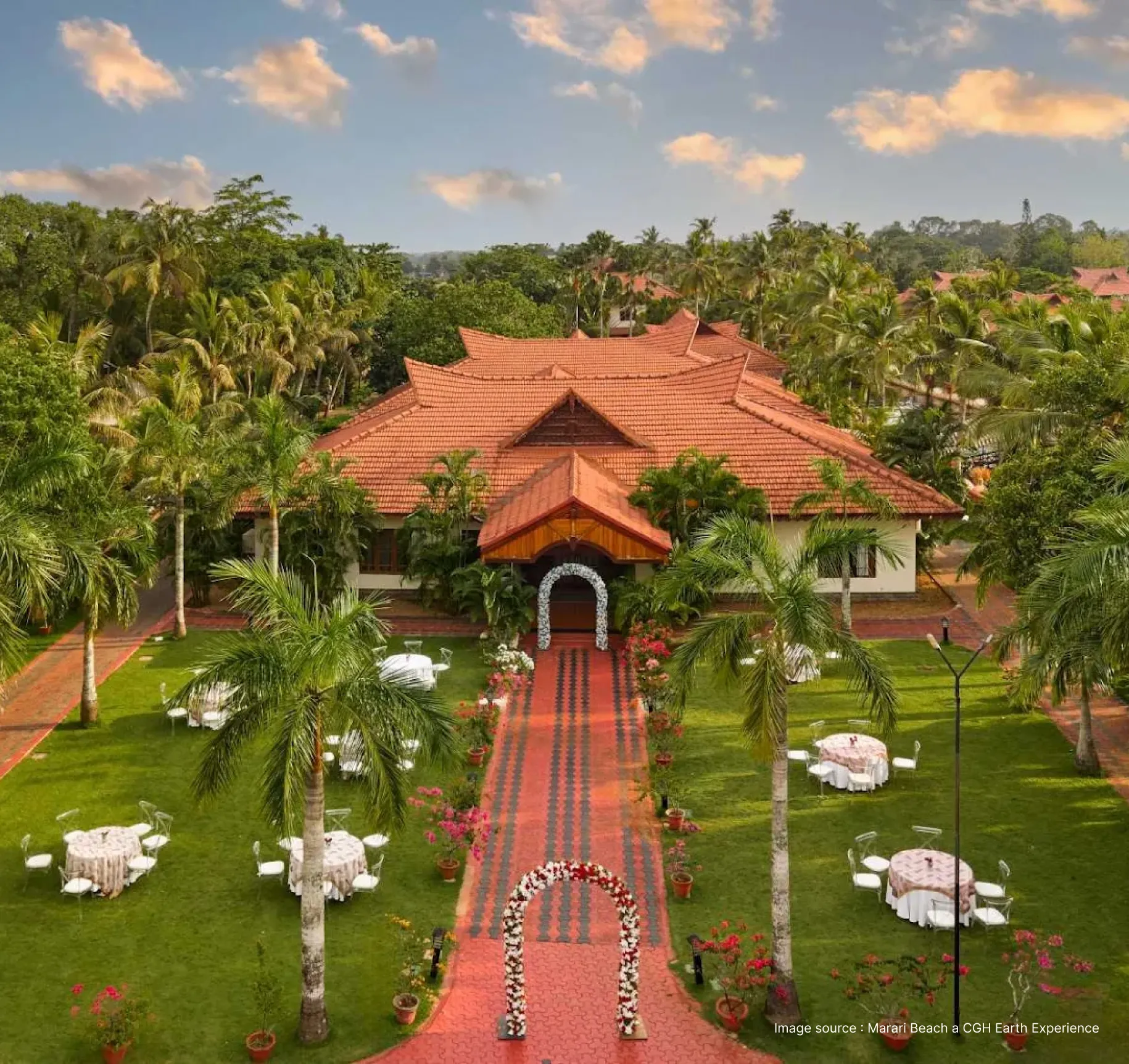 the grand entrance of the sterling lake palace in alleppey, surrounded by lush green palm trees & round tables for dining the garden